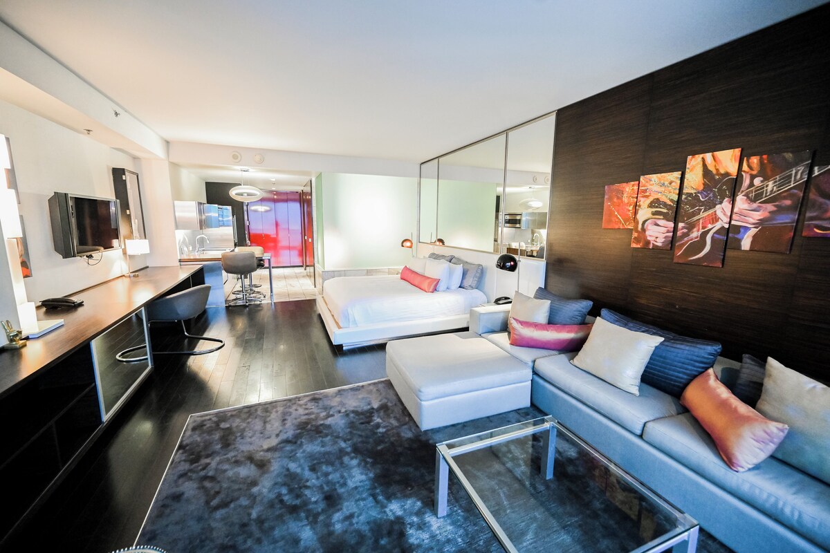 A modern hotel room with a bed, sofa, desk, and a large mirror.