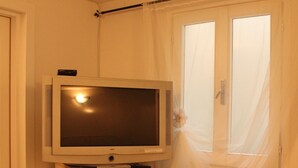 A compact room with a television on a cabinet, a window with sheer curtains, and a radiator.