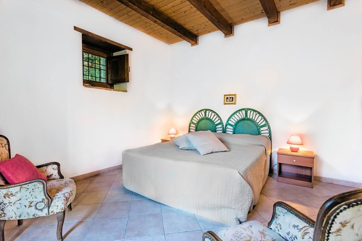 A bedroom with a bed, two nightstands, a chair, a small window, and a wooden ceiling.