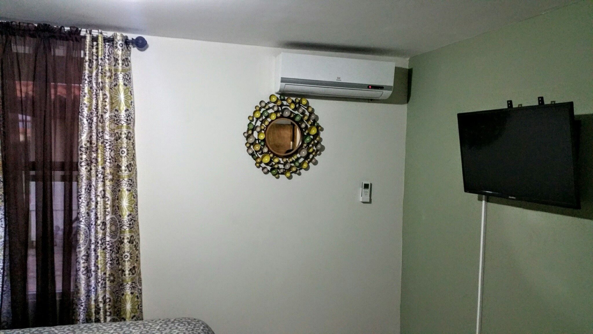 Habitación con cortina estampada, unidad de aire acondicionado, espejo decorativo, termostato y televisor montado.