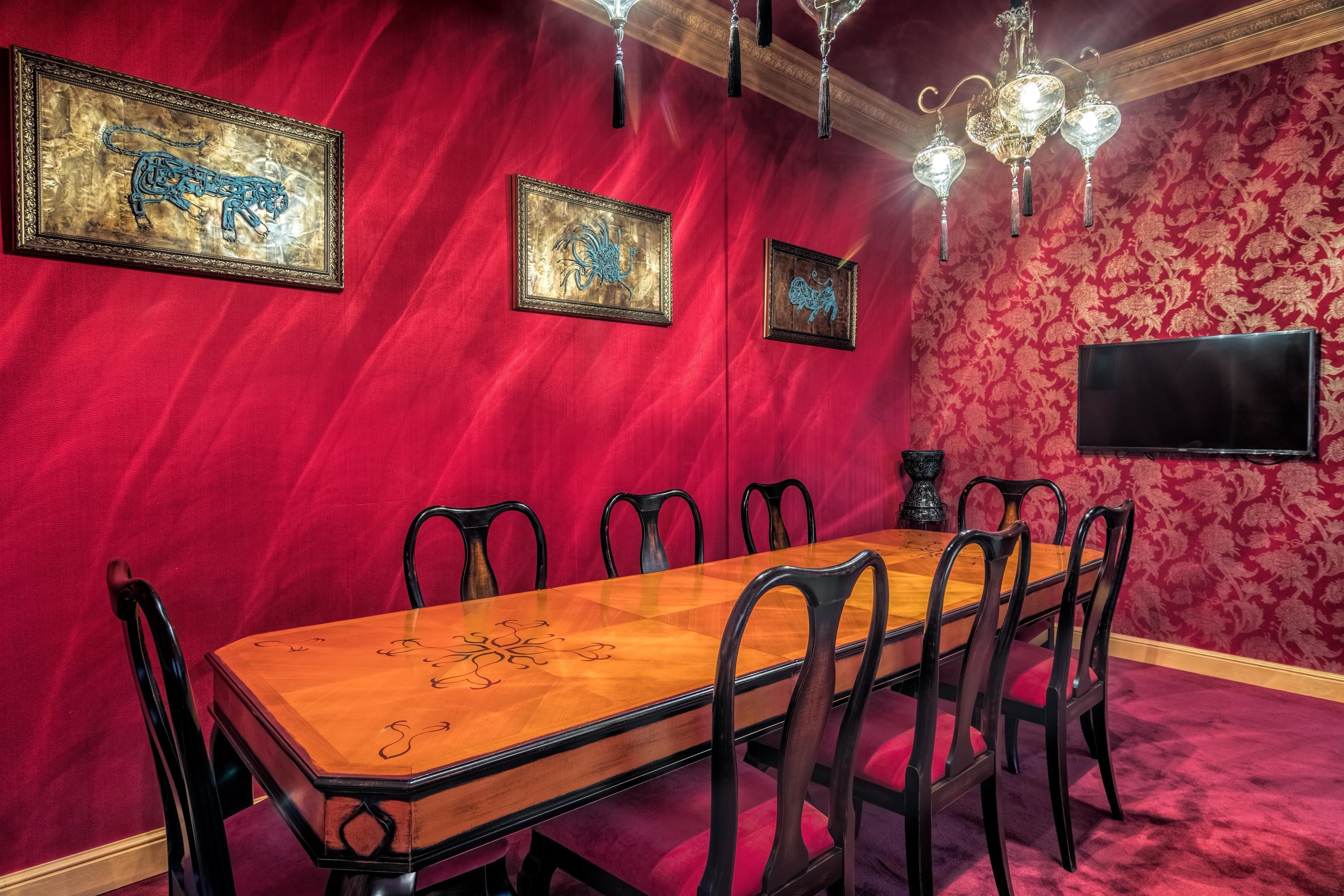 A room with a wooden dining table, six chairs, and red walls. There are three framed artworks on the wall and a chandelier.
