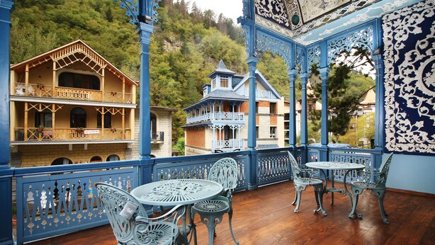 Un balcón con barandillas azules ornamentadas y sillas blancas, con vistas a una ladera boscosa.