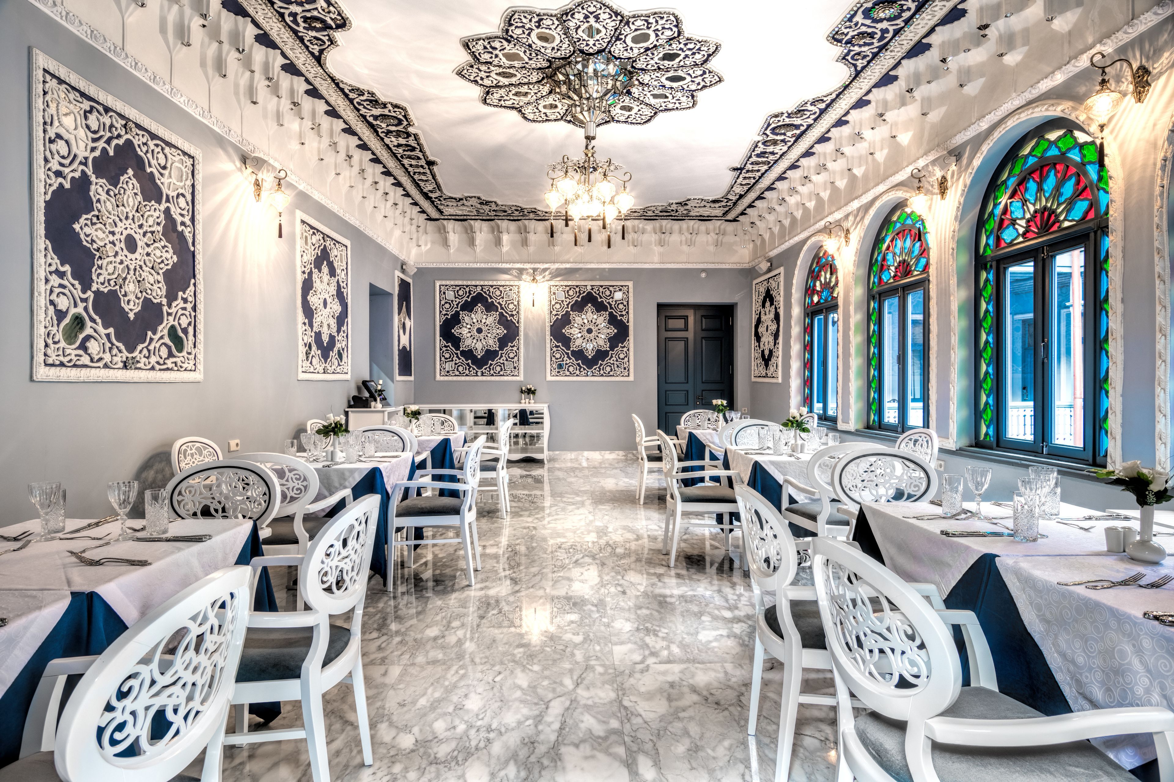 A dining area with white chairs, tables set with glassware, and intricate wall designs.