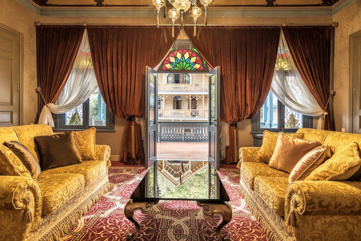 Una habitación lujosa con dos sofás ornamentados, una mesa de centro de vidrio y vistas a un balcón a través de una ventana con vitrales.