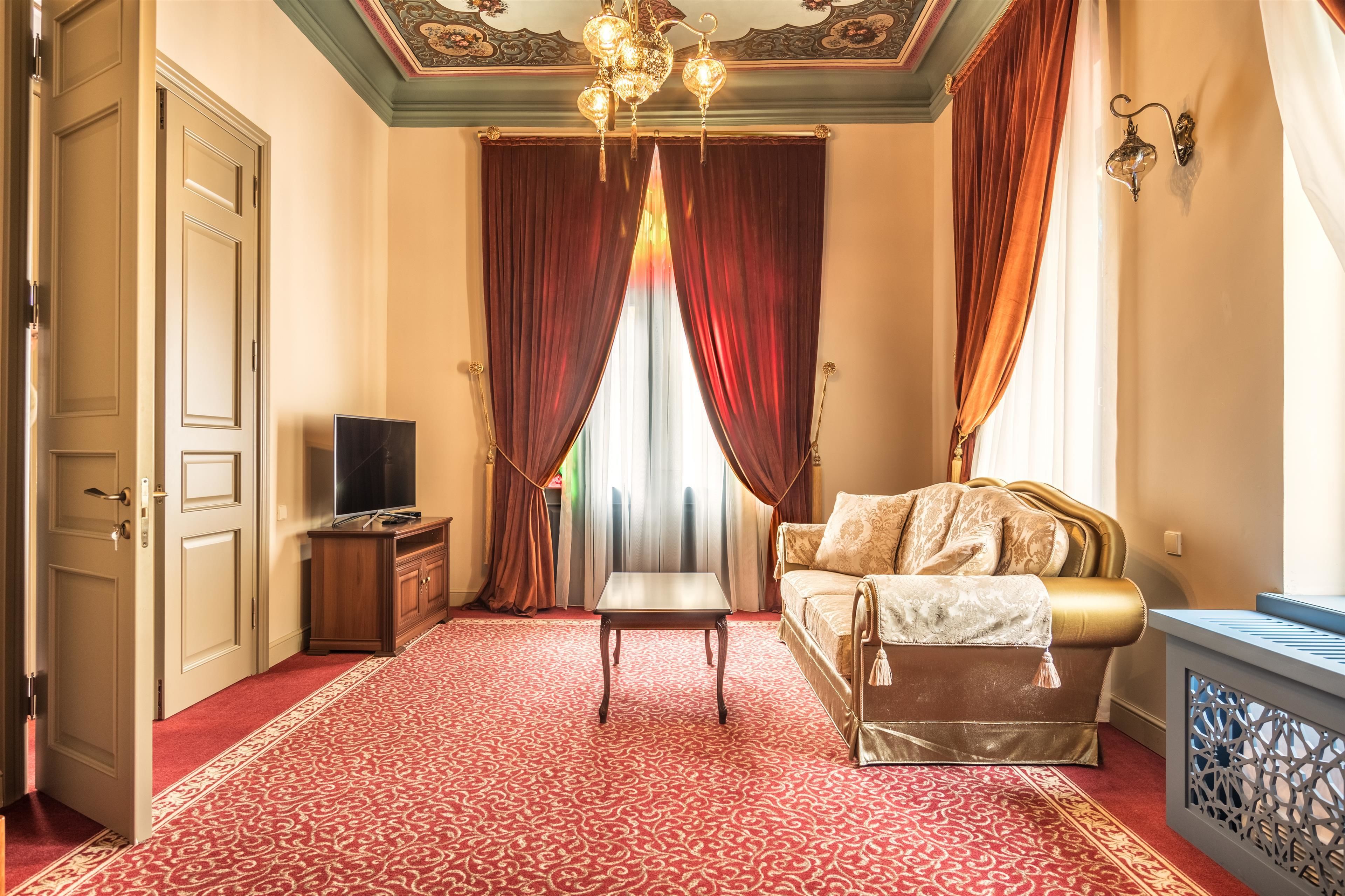 A room with a red patterned carpet, a bed with a gold headboard, a wooden nightstand, a television, and a chandelier.
