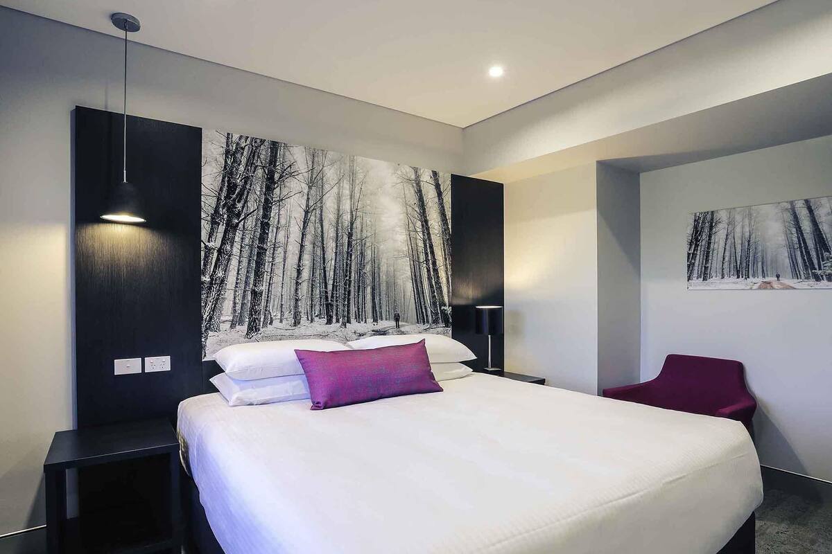 Een moderne hotelkamer met een groot bed, een nachtkastje, een rode stoel en een wanddecoratie waarop een boslandschap is afgebeeld.