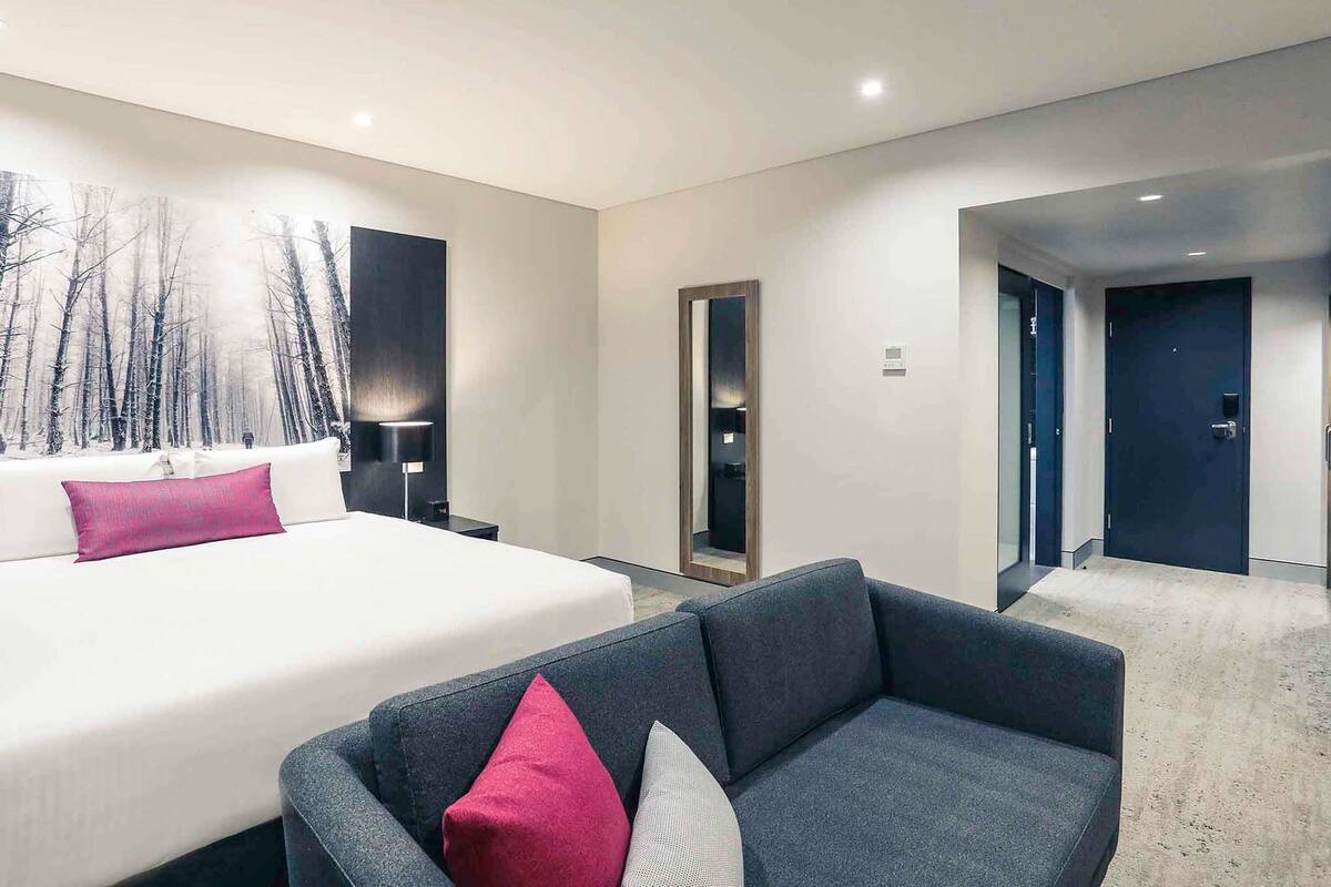 Een moderne hotelkamer met een groot bed, een donkere bank, een spiegel aan de muur en een deur naar een andere kamer.