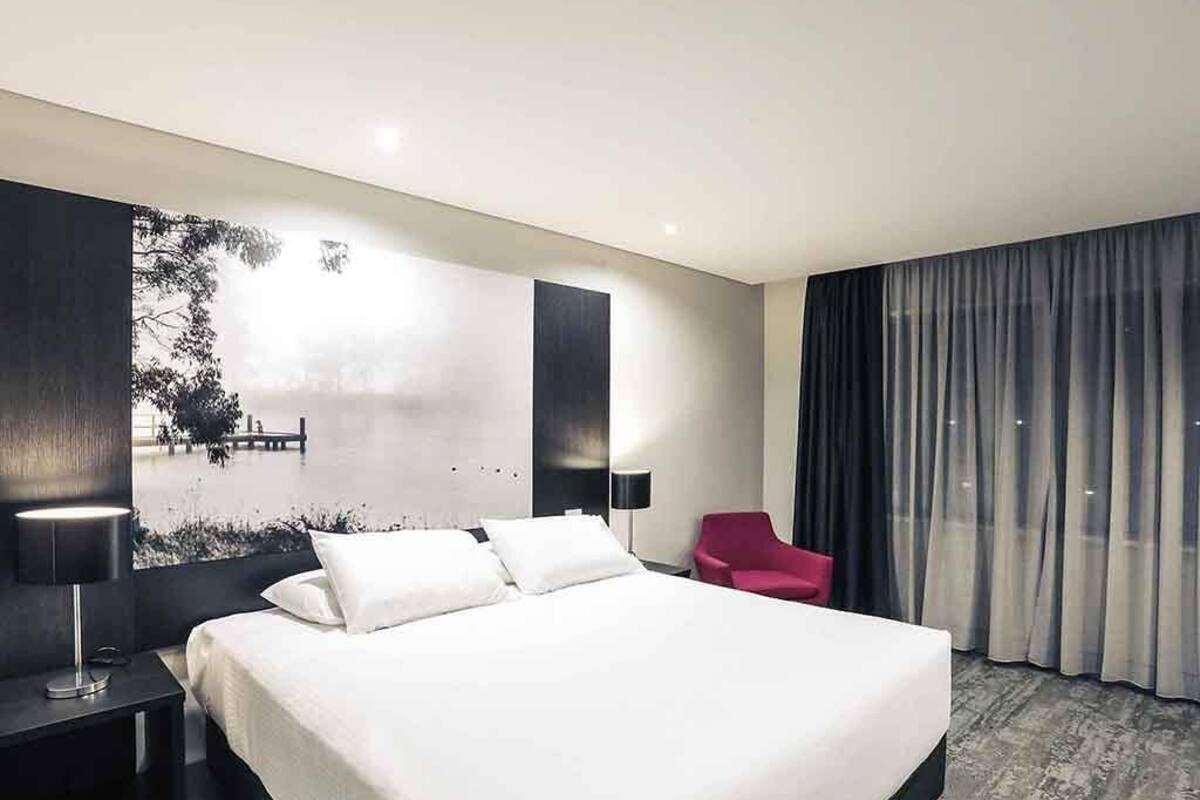 A modern hotel room with a large bed, a red chair, a wall-mounted TV, and a view of the outdoors.