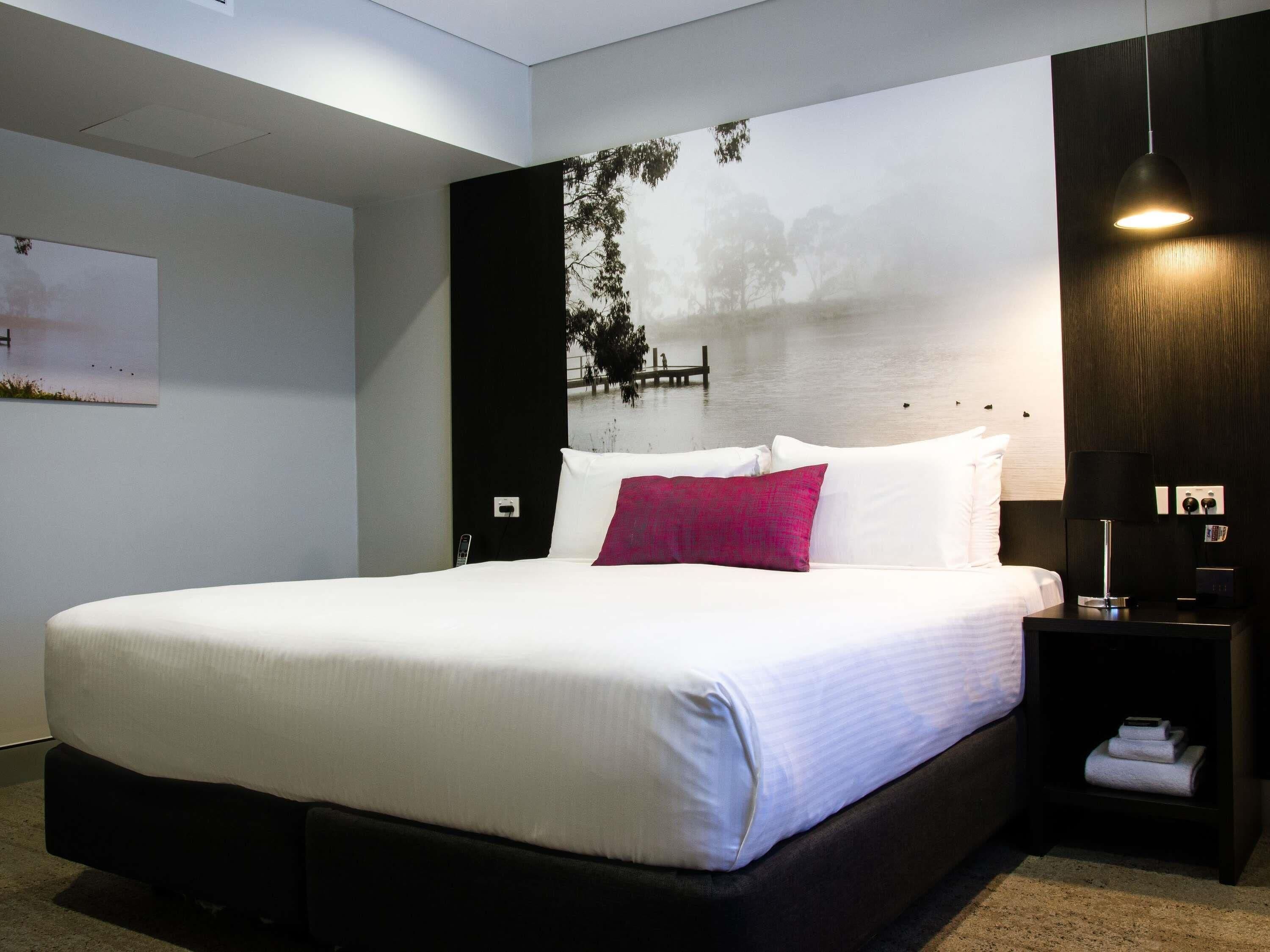 Ein modernes Hotelzimmer mit einem großen Bett, einem Nachttisch mit Lampe und einem Wandbild, das eine ruhige Landschaft zeigt.