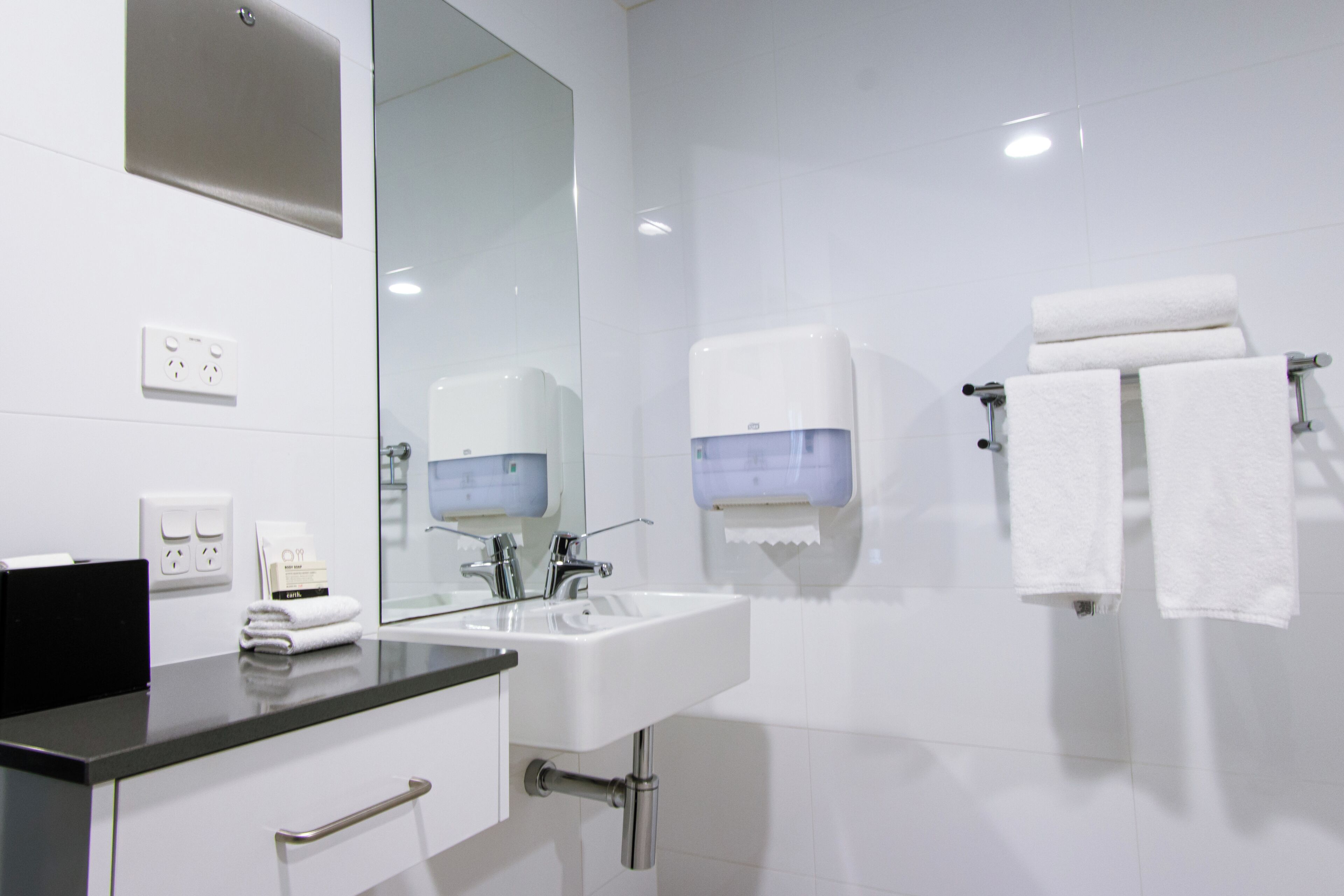A modern bathroom with a sink, mirror, towel dispenser, and a towel rack.