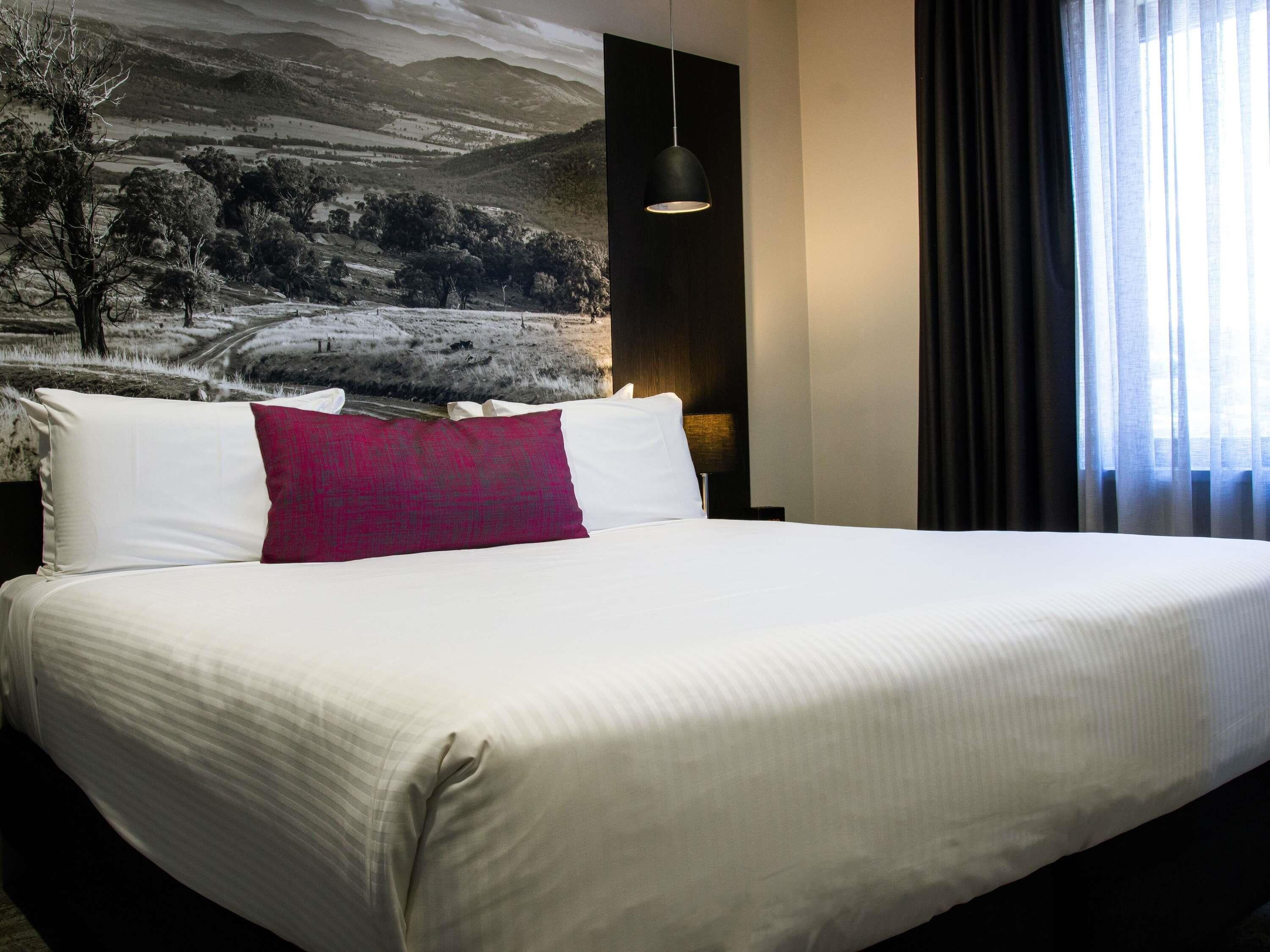 A hotel room with a large bed, a nightstand, a lamp, and a view of a landscape through the window.