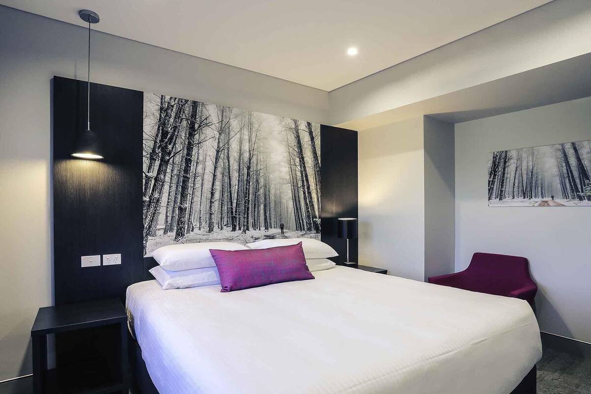 A modern bedroom with a large bed, a nightstand, a red chair, and a wall-mounted painting.