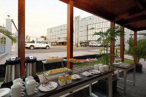 Mesa de buffet con diversos alimentos y máquinas de café, con vista a una calle con coches y un edificio al fondo.