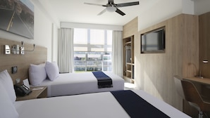 Habitación de hotel con dos camas, ventilador de techo, televisión, escritorio y ventana con vista a la ciudad.
