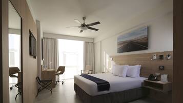 A hotel room with a large bed, a desk, two chairs, a ceiling fan, a painting, and a nightstand with a phone and a sign.