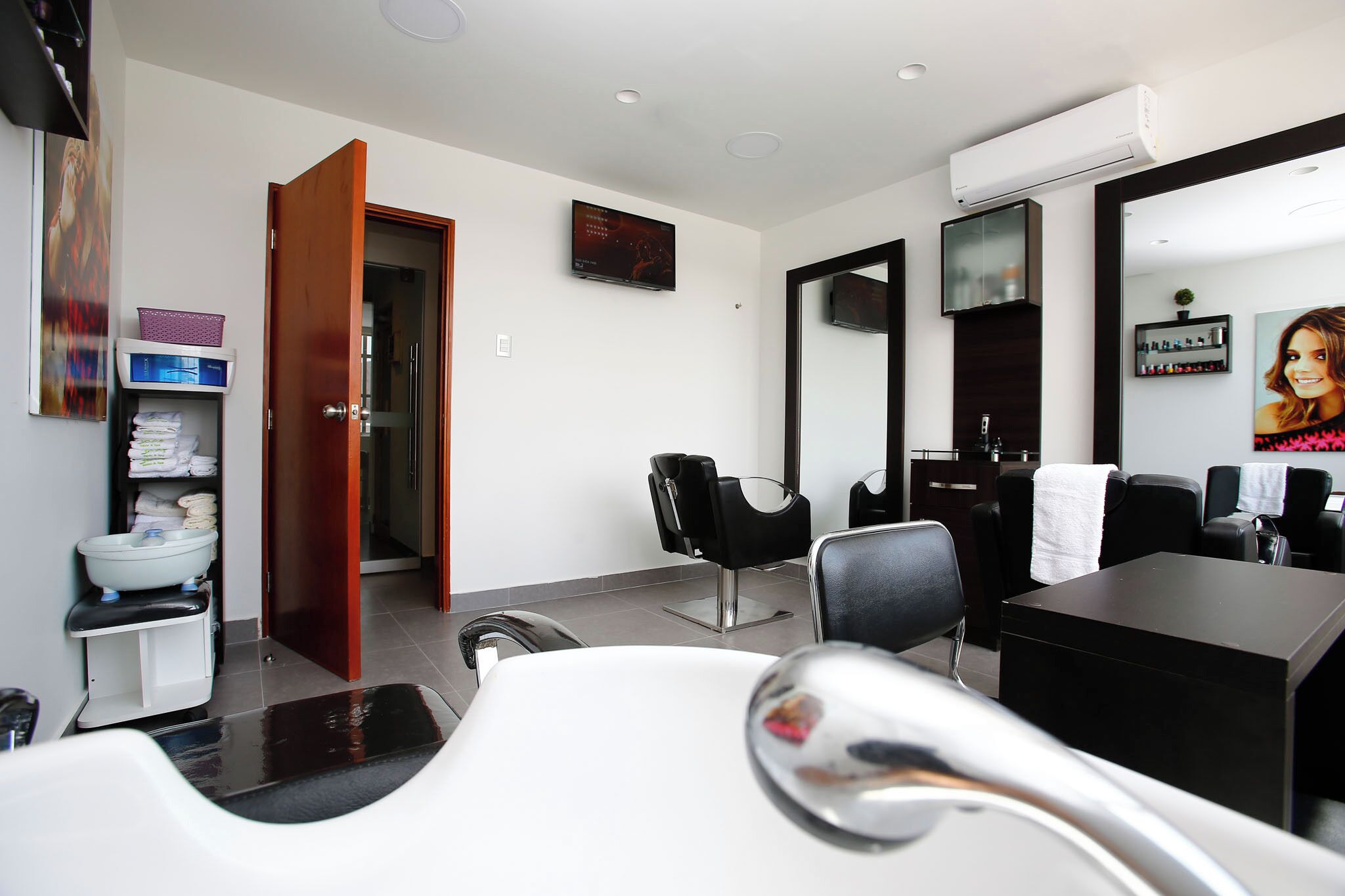 A modern hair salon with a reception desk, black salon chairs, a large mirror, and a television on the wall.