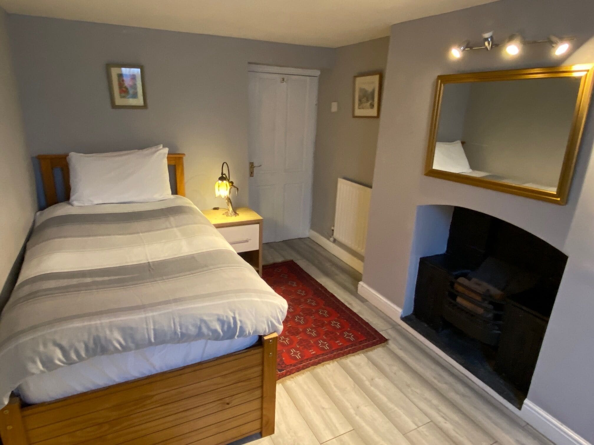 A bedroom with a bed, bedside lamp, dresser, mirror, fireplace, and a red rug.
