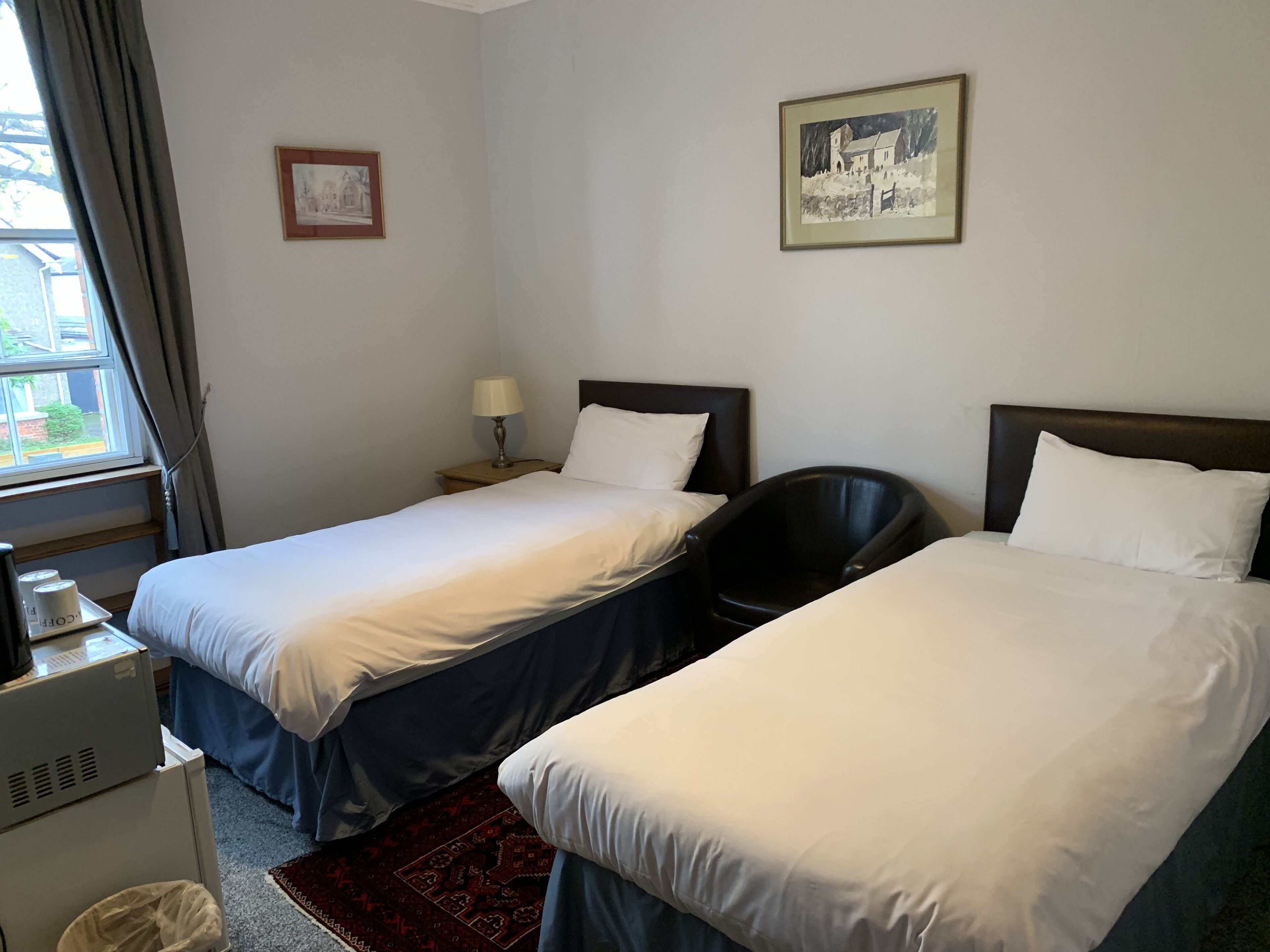 Een hotelkamer met twee eenpersoonsbedden, een bureau met lamp, een stoel en een raam met gordijnen.