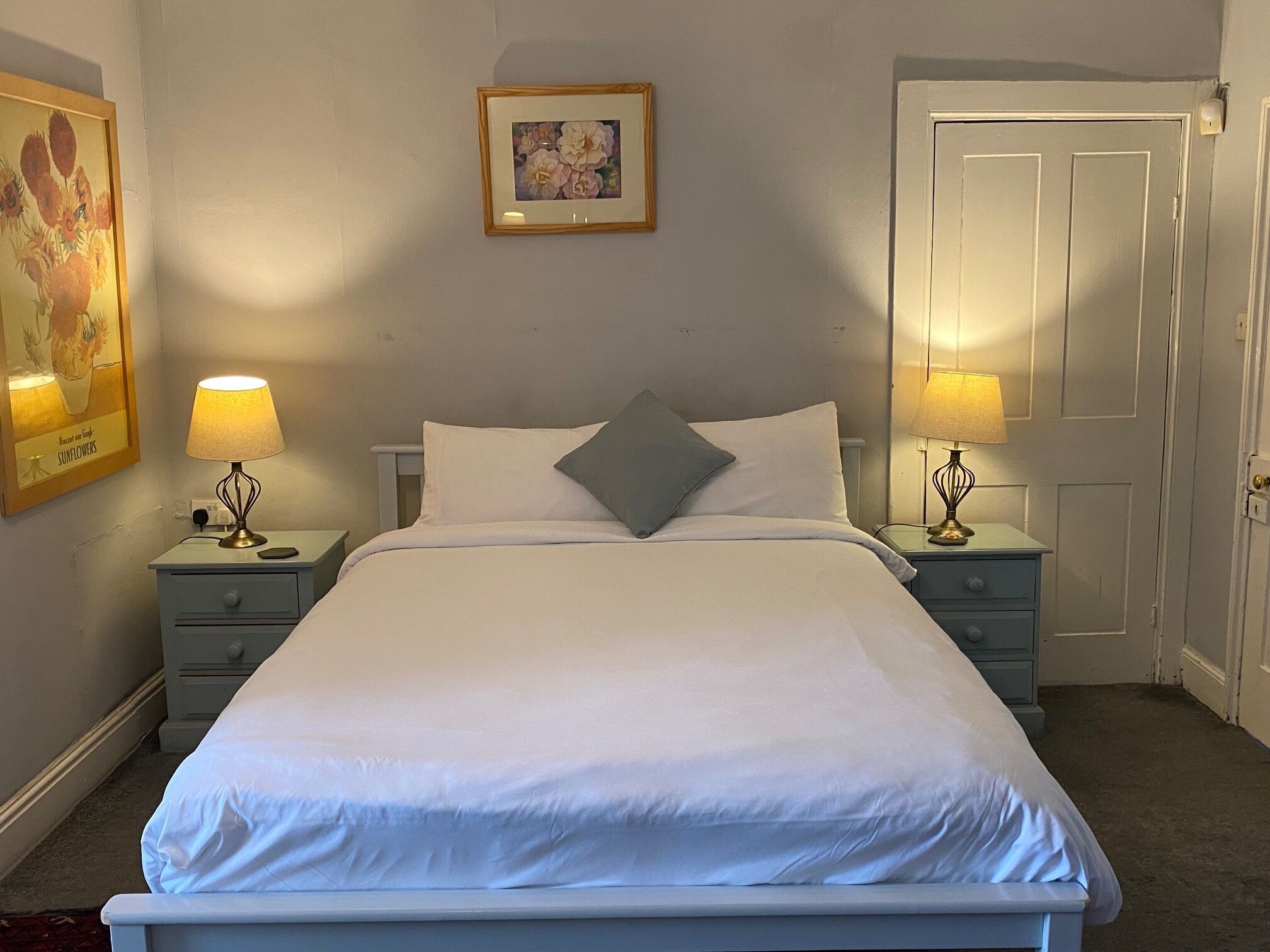 Una cama bien hecha con ropa blanca, dos mesitas de noche con lámparas, una puerta y un cuadro en la pared.