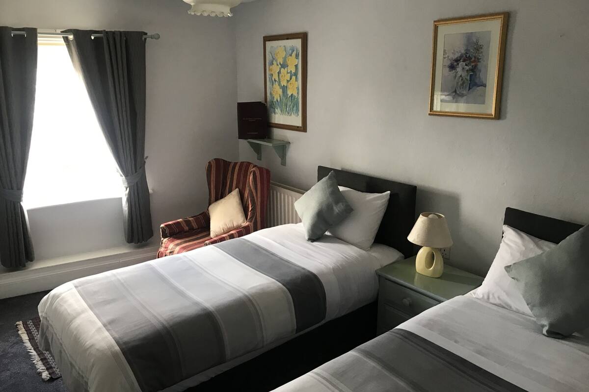 A hotel room with two beds, a chair, a nightstand with a lamp, and two framed pictures on the wall.