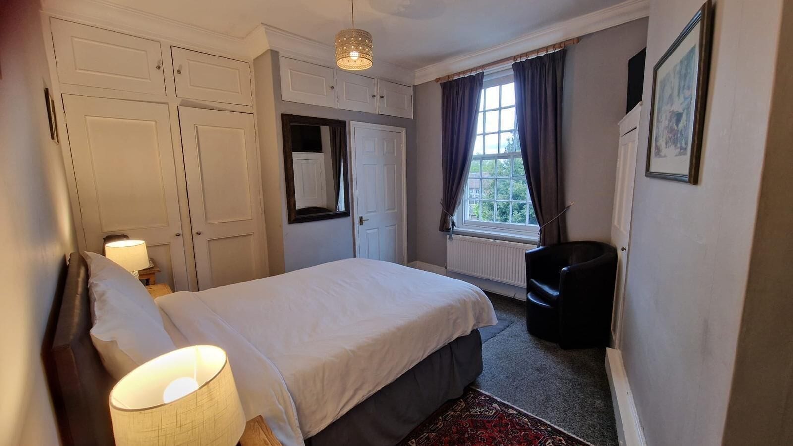 A bedroom with a bed, a chair, a window with curtains, a radiator, and a wardrobe.