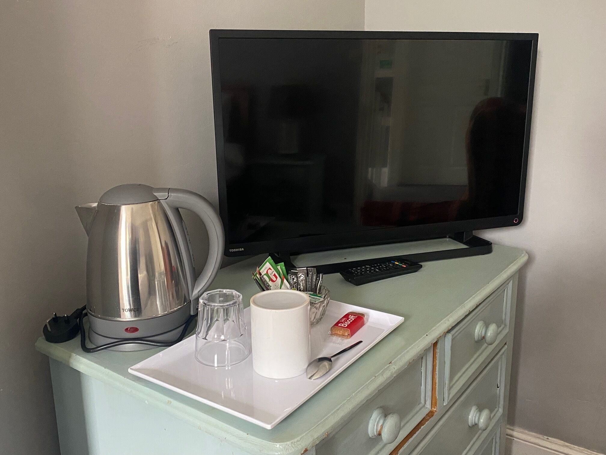 A kitchenette with a coffee maker, a TV, a remote control, and a plate with a mug and a spoon.