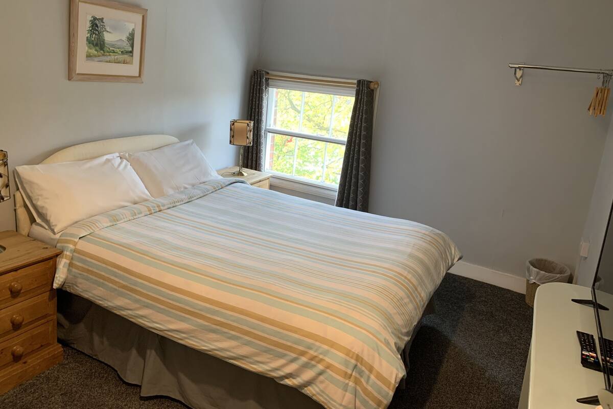 A neatly made bed with striped bedding, a bedside lamp, a wooden nightstand, a framed picture on the wall, and a window with curtains.