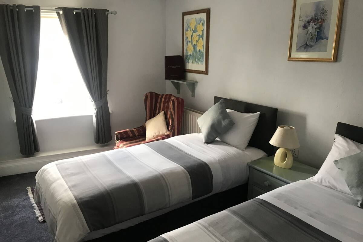 A hotel room with two beds, a chair, a nightstand with a lamp, and a painting on the wall.