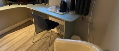 A room with a desk, a chair, a television, a window with curtains, and a white chair.