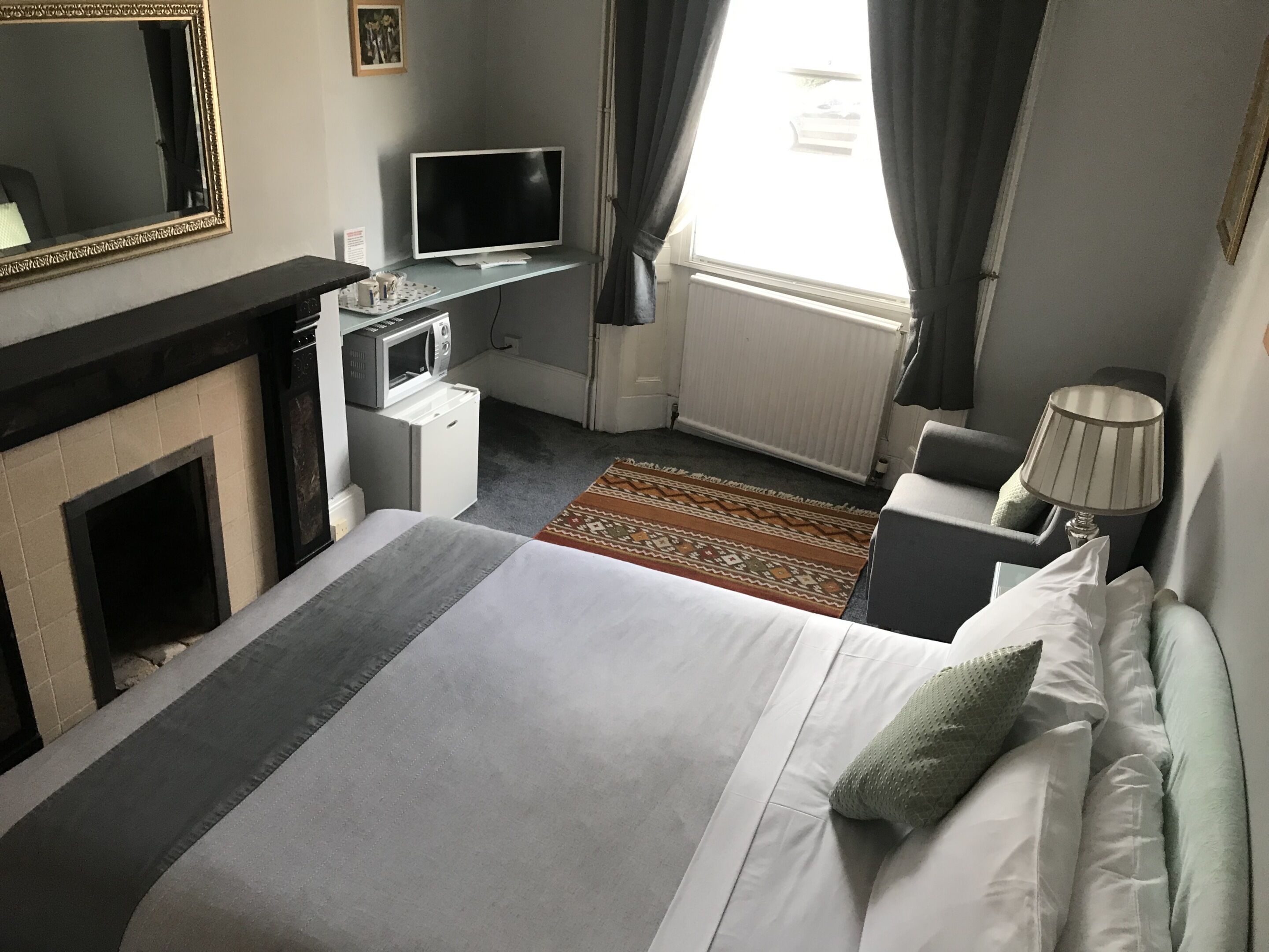 A hotel room with a bed, a desk with a computer, a microwave, a fireplace, and a window with curtains.