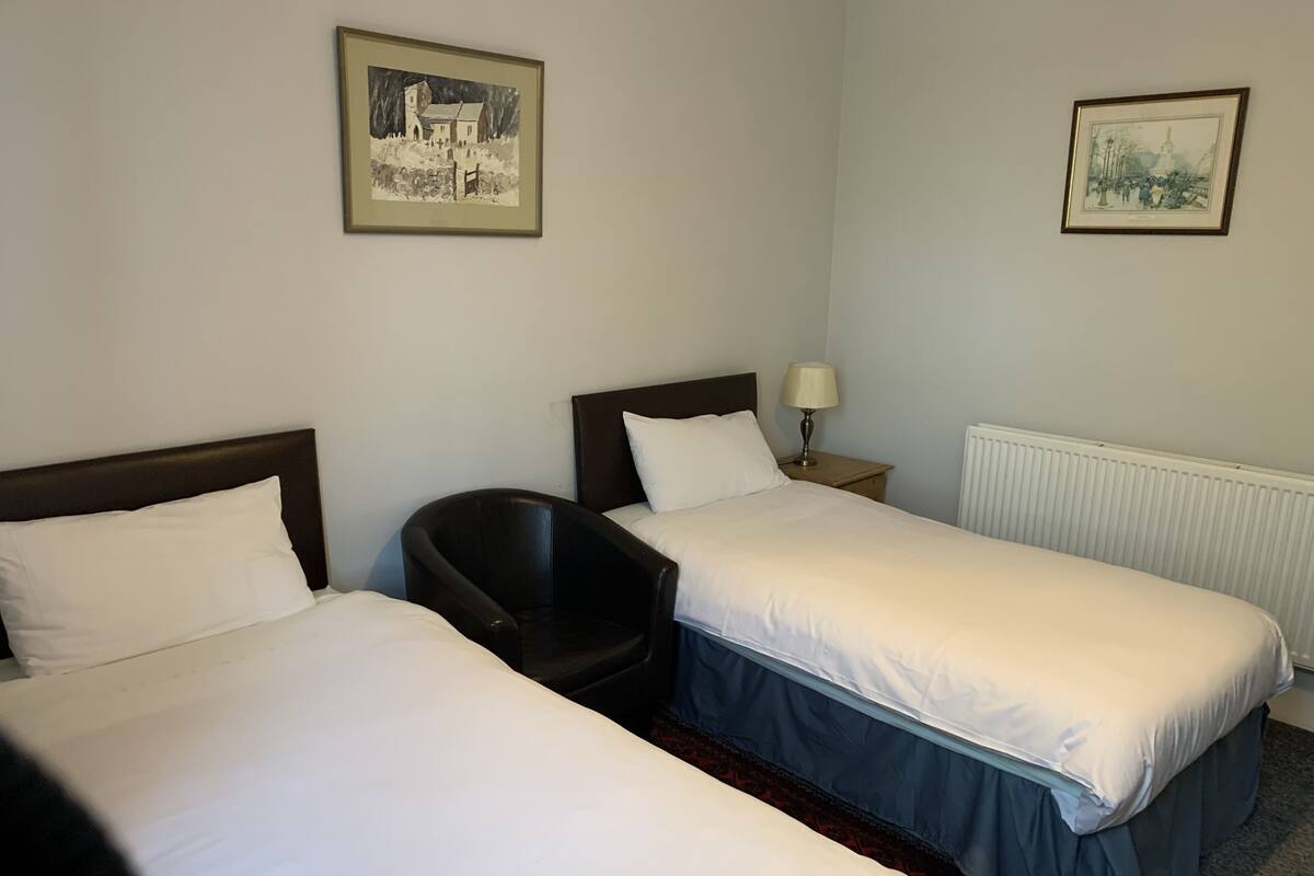 A hotel room with two single beds, a chair, a nightstand with a lamp, and two framed pictures on the walls.