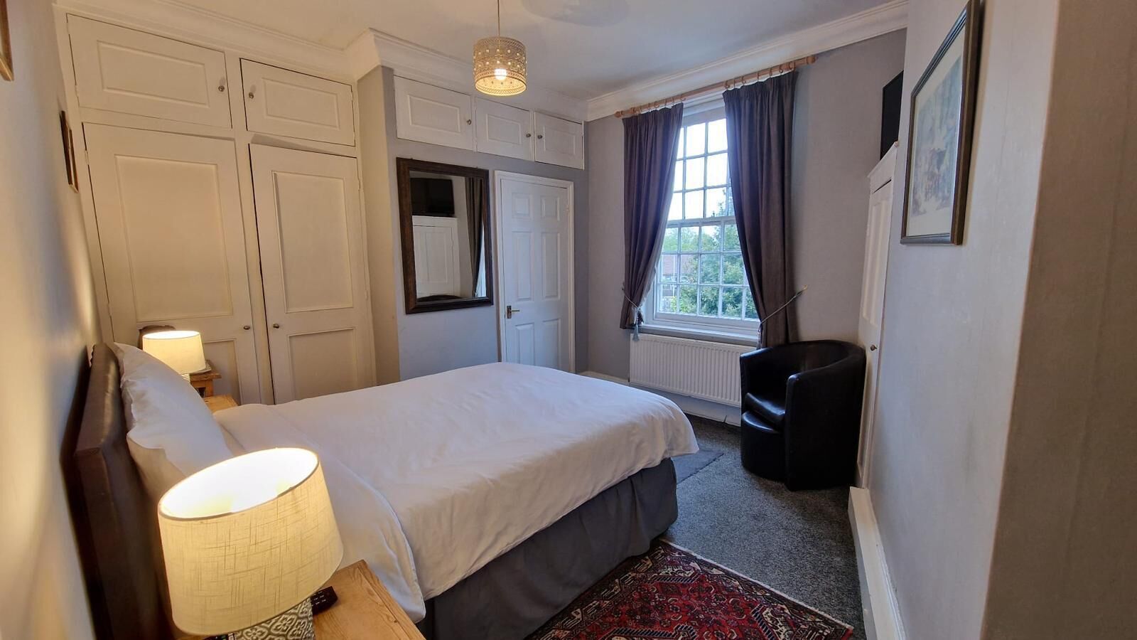 Une chambre à coucher comprenant un lit, une lampe de chevet, une armoire, une chaise, une fenêtre avec des rideaux et un radiateur.