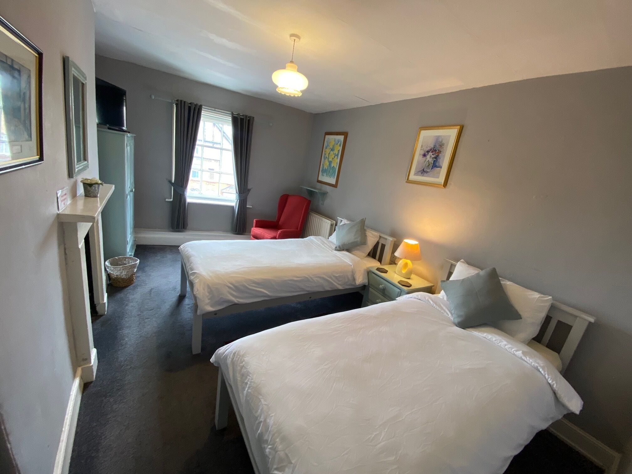 A hotel room with two beds, a red chair, a lamp, and a window with curtains.