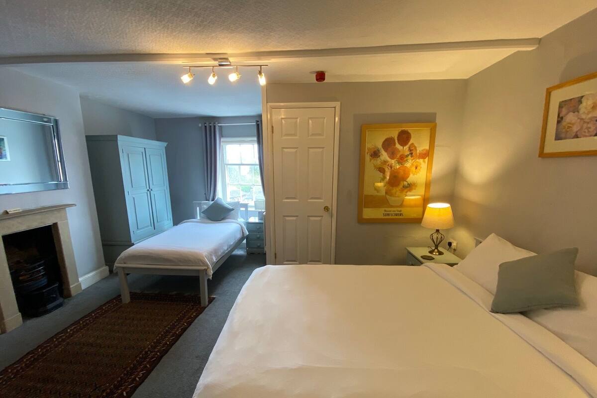 A hotel room with a large bed, a bedside table with a lamp, a small table with a lamp, a wardrobe, and a fireplace.