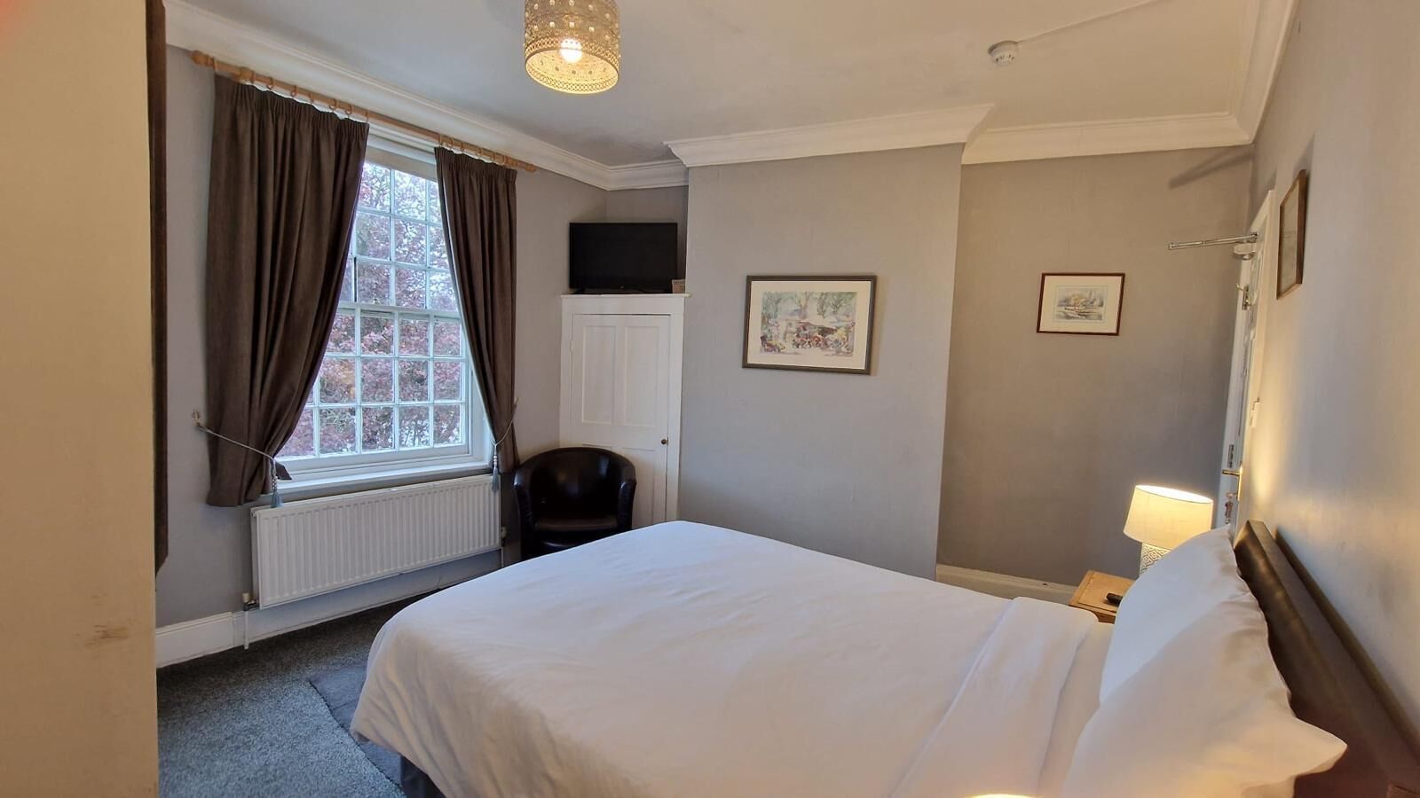 A hotel room with a bed, a chair, a radiator, a window with curtains, and a wall-mounted picture.