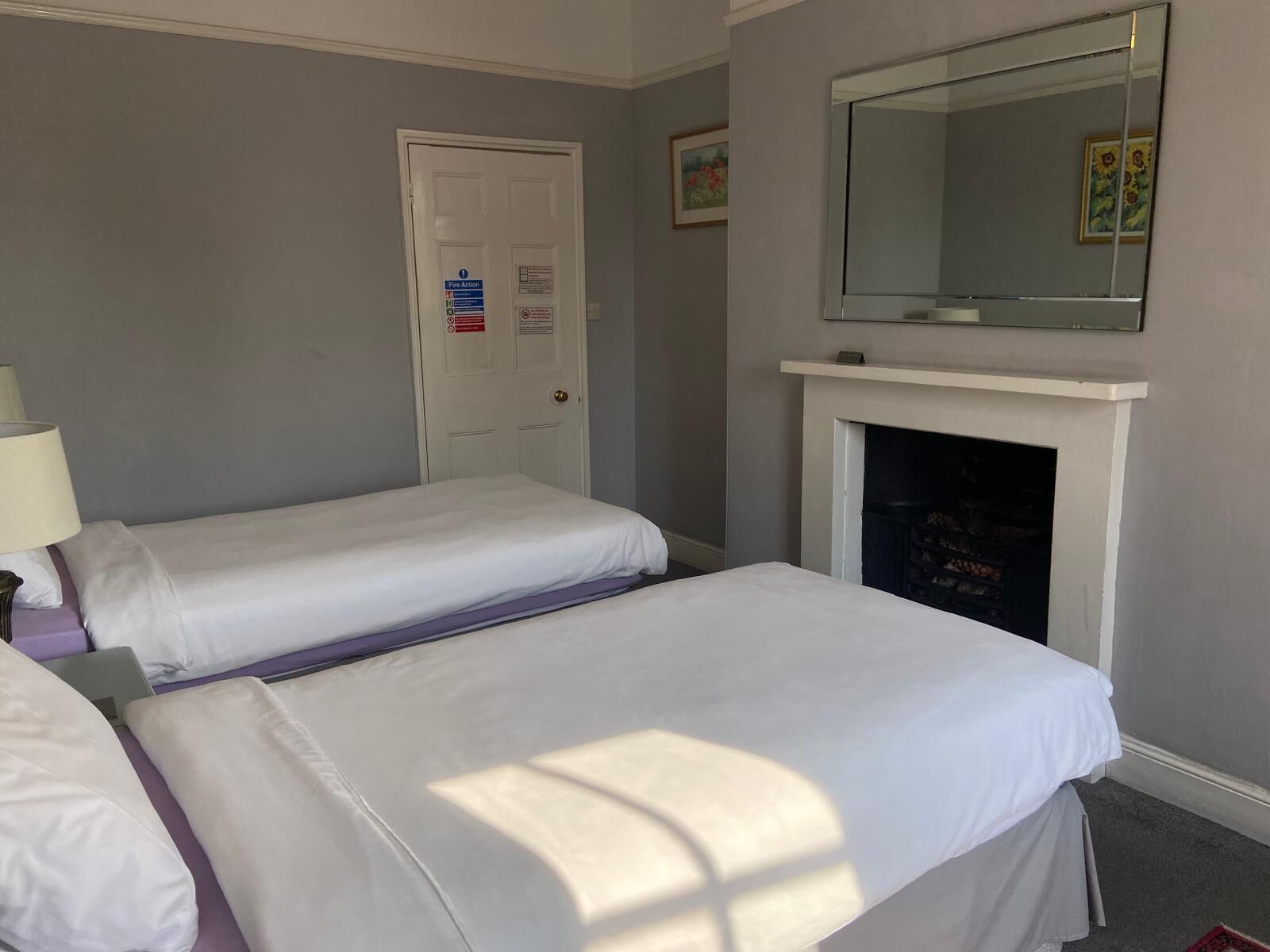A hotel room with two beds, a white door with fire action instructions, a fireplace, and a mirror above it.
