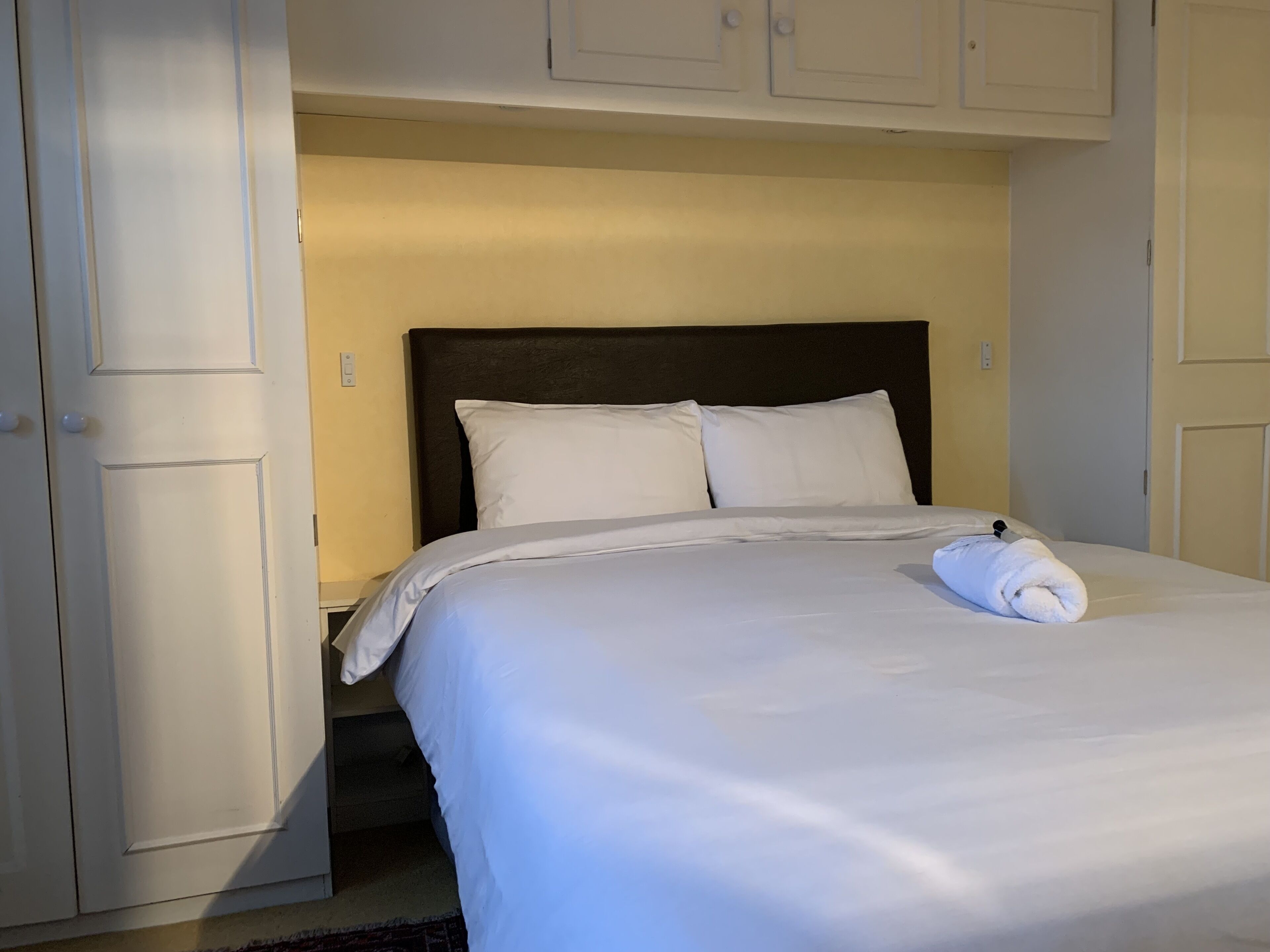 Una cama bien hecha con sábanas y almohadas blancas, cabecera oscura y una toalla enrollada al pie.