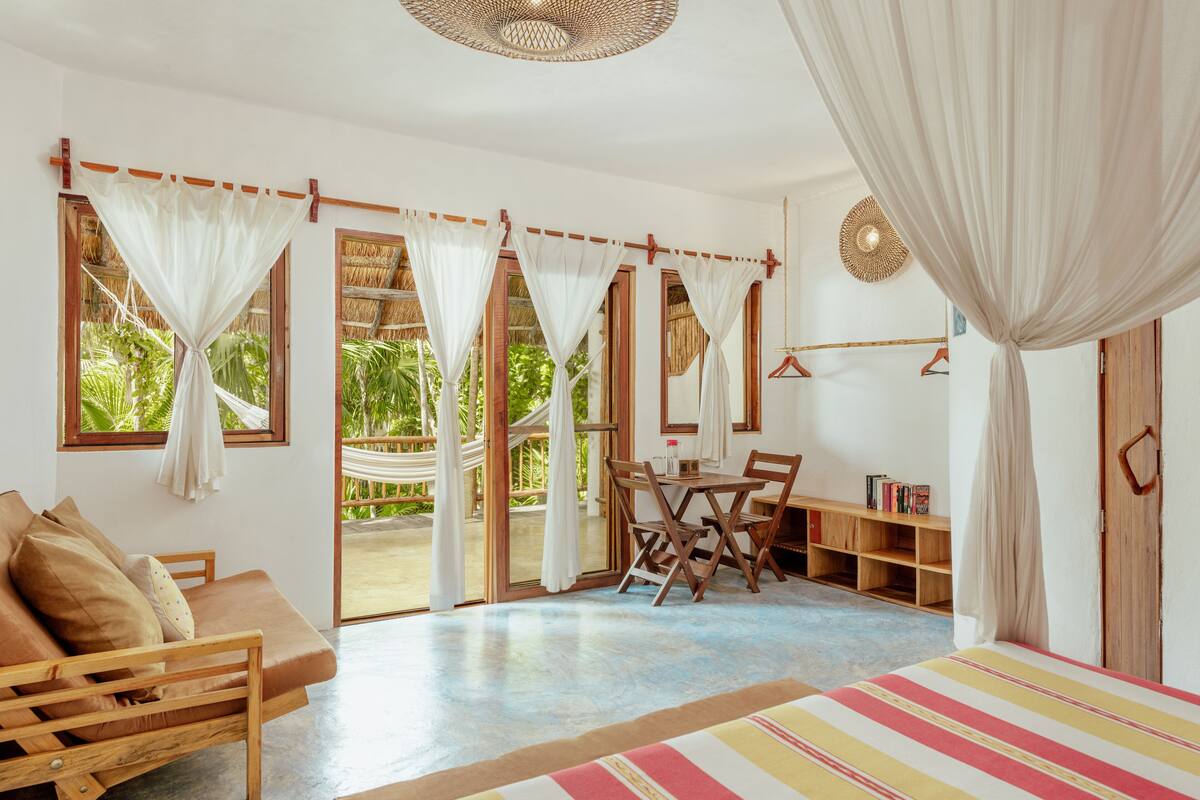 A room with a wooden sofa, a striped bed, a dining table with chairs, a bookshelf, and a hammock.