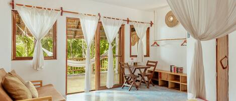 A room with a wooden sofa, a striped bed, a dining table with chairs, a bookshelf, and a hammock.