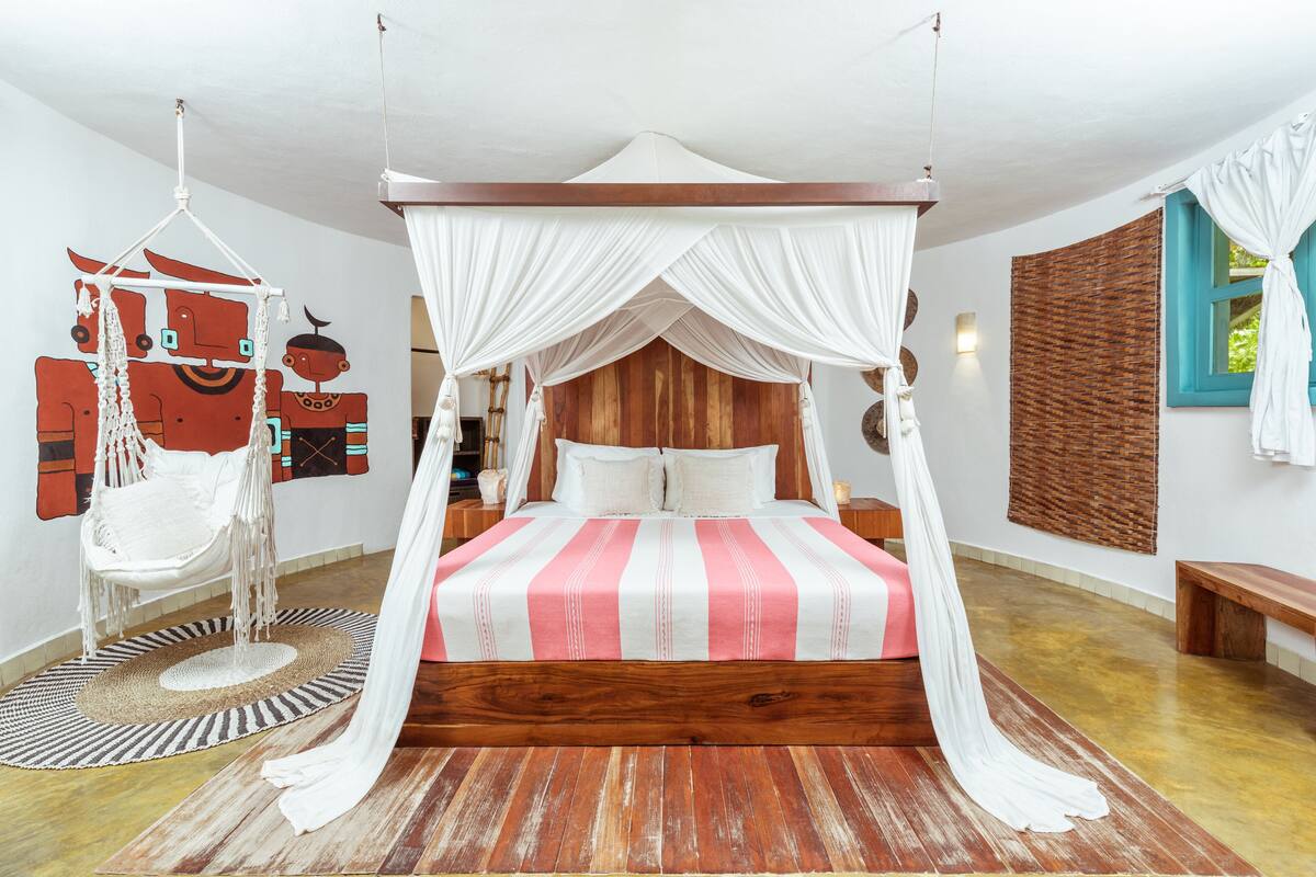 A four-poster bed with a striped bedspread, a hanging chair, and a woven wall hanging.
