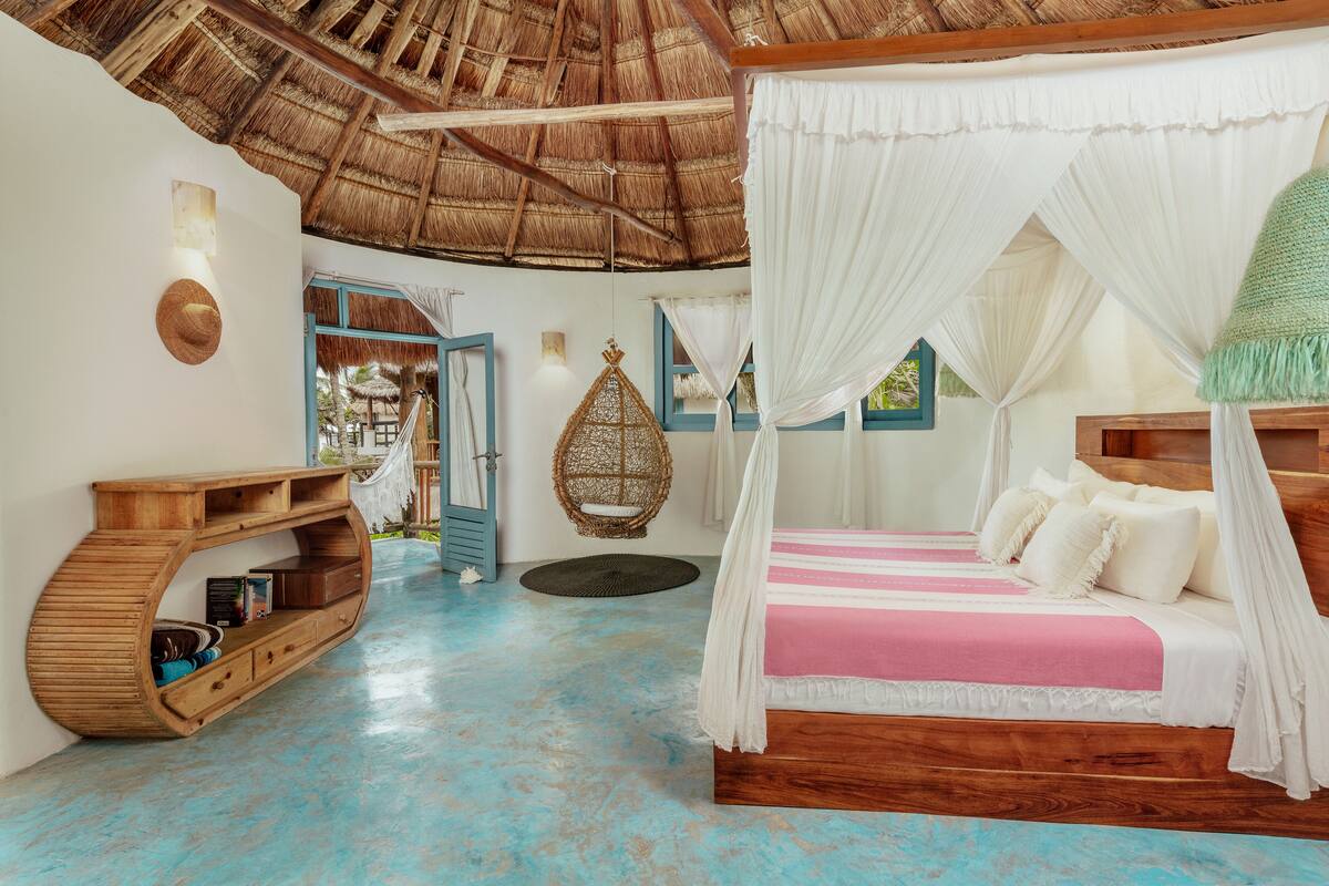 A room with a bed, a wooden console table, a hanging chair, and a thatched ceiling.