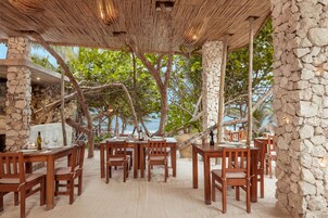 Un área de comedor al aire libre con muros de piedra, mesas y sillas de madera, rodeada de árboles y vistas a una playa.