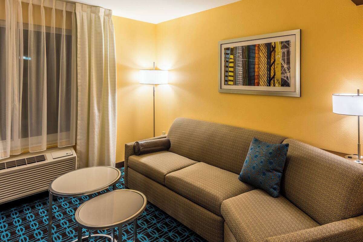 Une chambre d’hôtel avec un canapé, deux petites tables, une lampe, un tableau au mur et une fenêtre avec des rideaux.