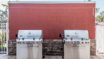 Twee Weber barbecues met meerdere branders, voor een rode muur met een stenen onderbouw, op een houten terras.