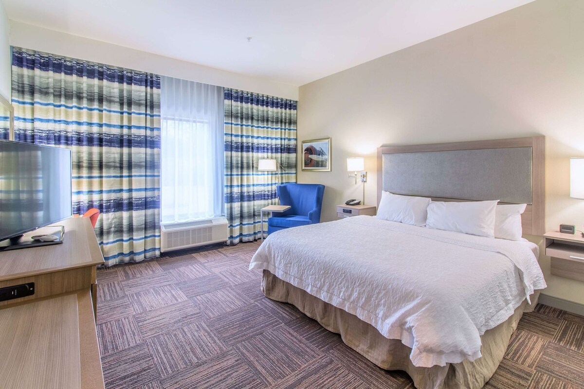 A hotel room with a large bed, a desk with a TV, a chair, a bedside table with a lamp, and a window with curtains.