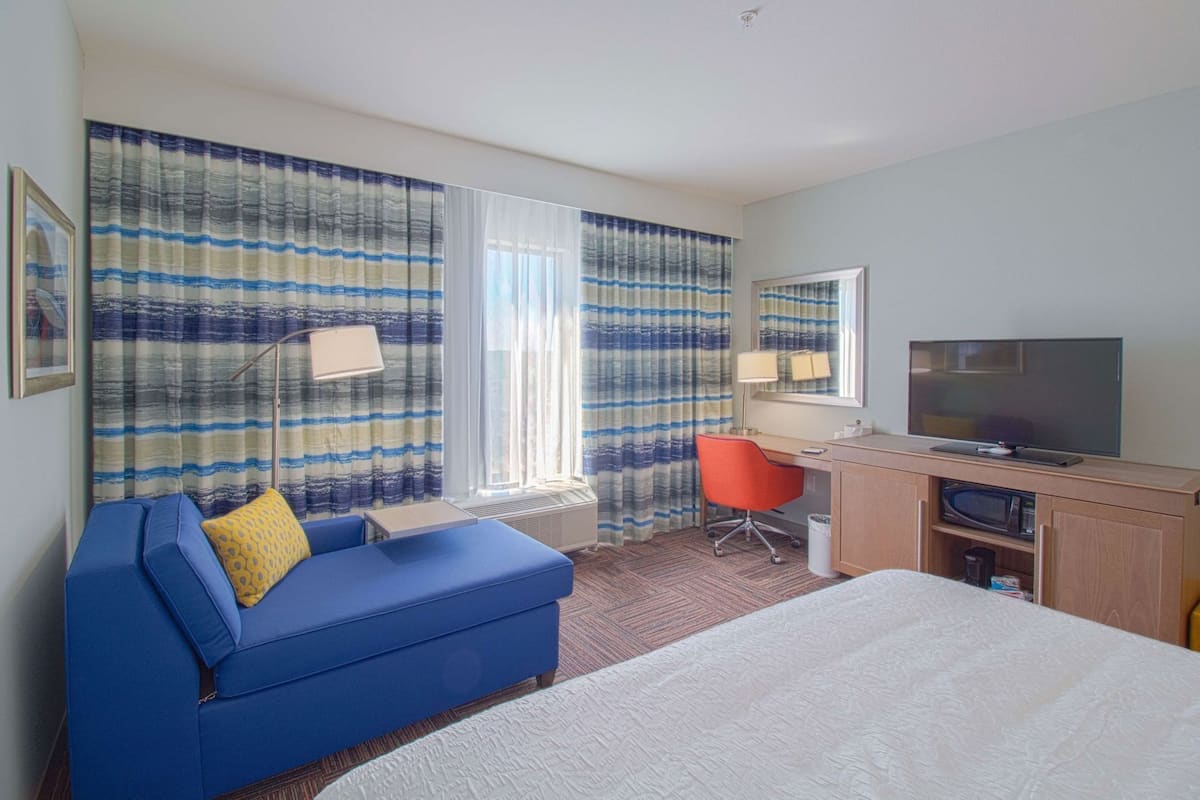 A hotel room with a bed, a blue sofa, a desk with a chair, a television, and curtains.
