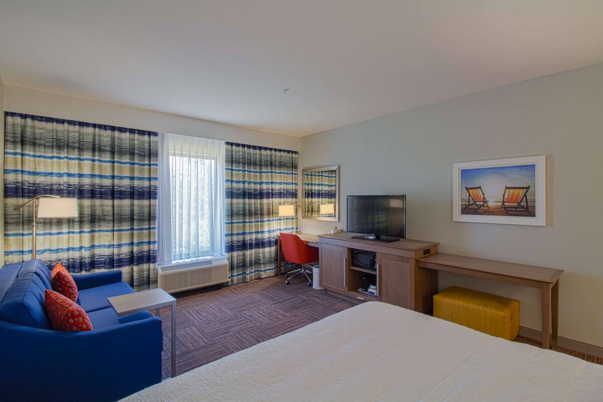 A hotel room with a bed, a desk with a computer, a TV, a red chair, and a window with curtains.