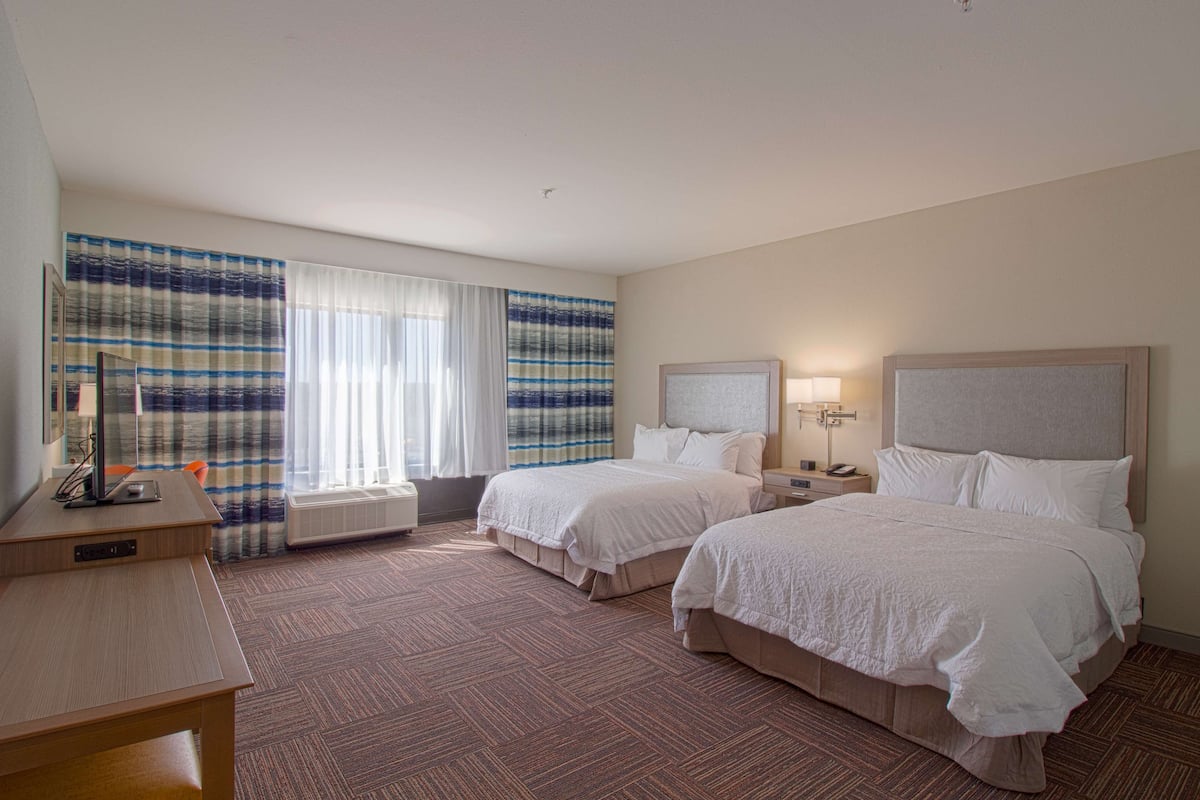 A hotel room with two beds, a desk, a chair, a nightstand with a lamp, and a large window with curtains.