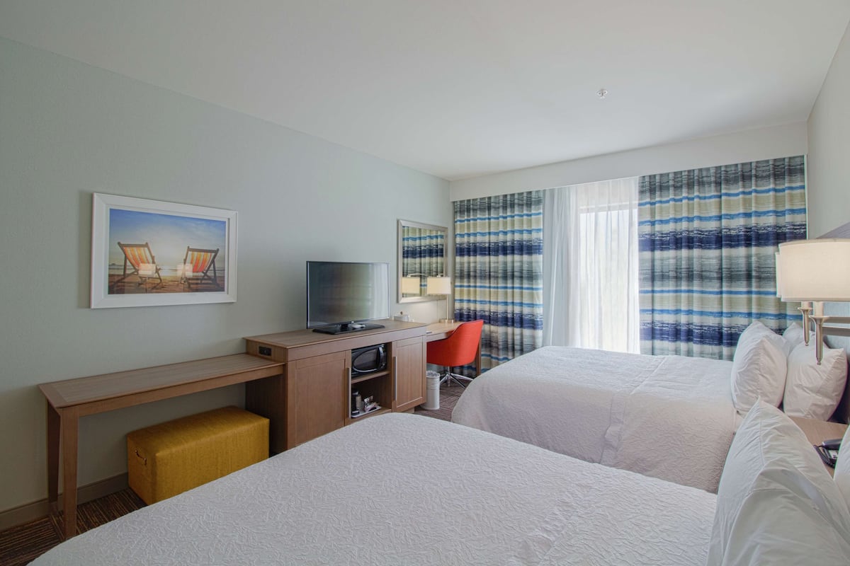 A hotel room with two beds, a desk with a television, a red chair, and a large window with striped curtains.