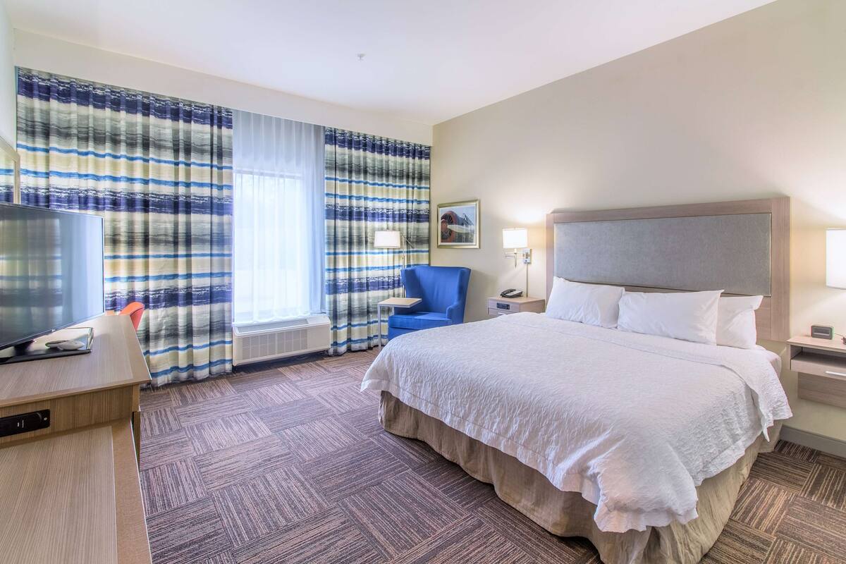 A hotel room with a large bed, a desk with a television, a chair, and a window with striped curtains.